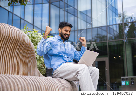 A man celebrates success while working outdoors on a laptop, showcasing modern business and achievement. A man celebrates success while working outdoors on a laptop, showcasing modern business and achievement. 129497038
