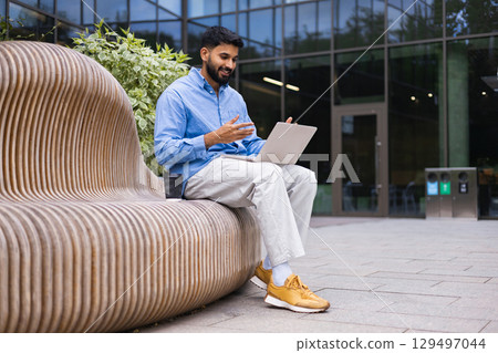 A businessman on a video call outside, sitting on a unique wooden bench with his laptop. A businessman on a video call outside, sitting on a unique wooden bench with his laptop. 129497044