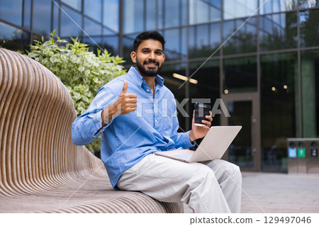 A man with a beard gives a thumbs up while holding a coffee and laptop in front of a modern building. A man with a beard gives a thumbs up while holding a coffee and laptop in front of a modern building. 129497046