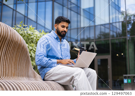 A man sits on a bench, using a laptop outdoors, with a coffee cup in hand, in front of a modern building. 129497063