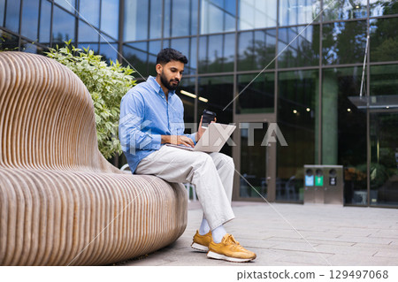 A man working on his laptop while sitting outside, holding a coffee cup. A man working on his laptop while sitting outside, holding a coffee cup. 129497068