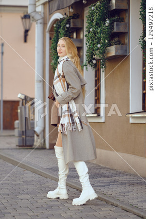 Woman in beige coat and white boots walks along a charming street adorned with greenery 129497218