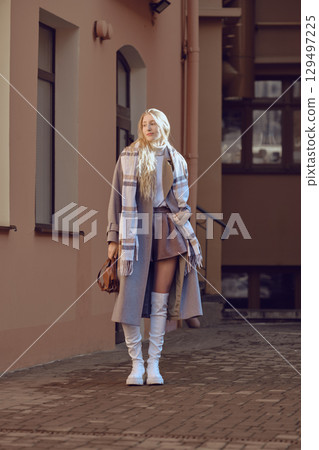 A fashionable young woman in wool coat, knee-high boots and scarf posing in a sunny urban alley 129497225