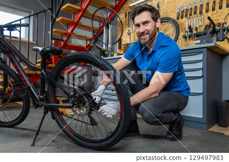 Bicycle mechanic in blue shirt kneeling with gloves adjusting cycle tire in organized indoor garage Bicycle mechanic in blue shirt kneeling with gloves adjusting cycle tire in organized indoor garage 129497983