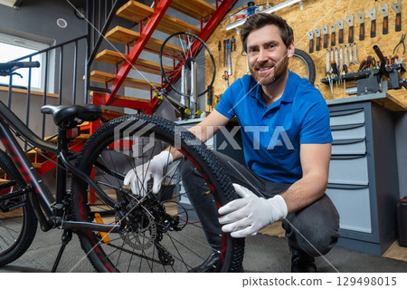 Smiling bearded man in blue polo shirt repairing bicycle wheel in modern indoor workshop Smiling bearded man in blue polo shirt repairing bicycle wheel in modern indoor workshop 129498015