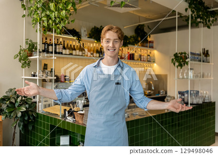 Cheerful male barista with apron smiling and welcoming guests with open arms standing behind counter Cheerful male barista with apron smiling and welcoming guests with open arms standing behind counter 129498046