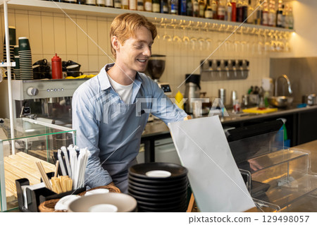 Pleased barista leaning slightly forward on bar counter smiling toothily standing inside coffee shop 129498057