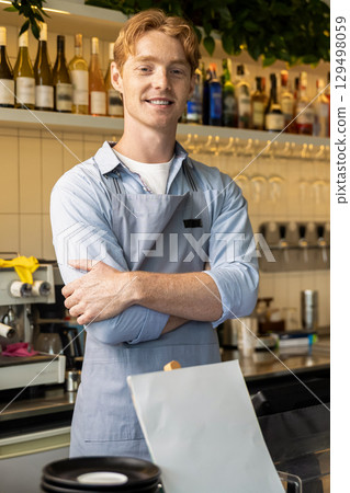 Confident male barista standing with arms crossed in front of counter wearing apron 129498059
