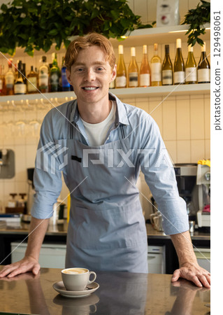 Joyful café employee in apron posing near coffee counter with hands on surface 129498061