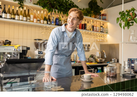 Smiling male coffee shop worker in apron standing at counter near espresso machine Smiling male coffee shop worker in apron standing at counter near espresso machine 129498062