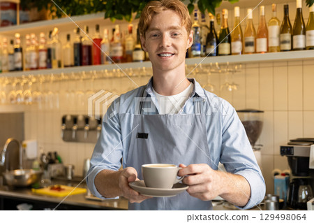 Smiling male barista in apron holding cup standing at counter in modern coffee shop interior 129498064