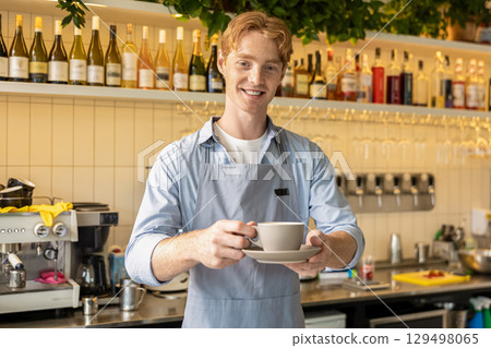 Cheerful male barista in apron standing behind coffee counter holding cup smiling at customer 129498065