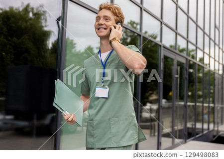 Ginger doctor standing by glass clinic building holding and discussing on smartphone Ginger doctor standing by glass clinic building holding and discussing on smartphone 129498083