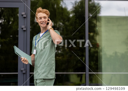 Doctor in green scrubs standing by hospital wall holding folder and discussing medicine on phone 129498109
