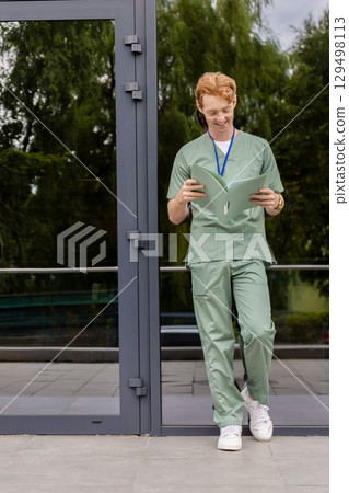 Caucasian doctor in green uniform standing near glass clinic facade with badge and folder 129498113
