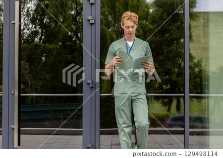 Red haired male medical professional standing near modern glass hospital carrying folder 129498114