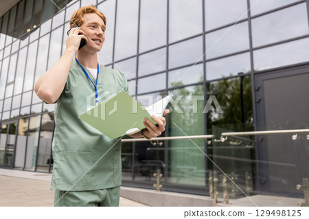 Ginger doctor standing by glass clinic building holding and discussing on smartphone Ginger doctor standing by glass clinic building holding and discussing on smartphone 129498125