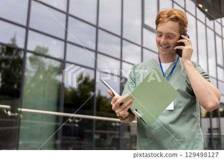 Young male healthcare professional in scrubs talking on smartphone near hospital Young male healthcare professional in scrubs talking on smartphone near hospital 129498127