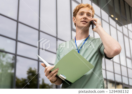 Young male healthcare professional in scrubs talking on smartphone near hospital Young male healthcare professional in scrubs talking on smartphone near hospital 129498132