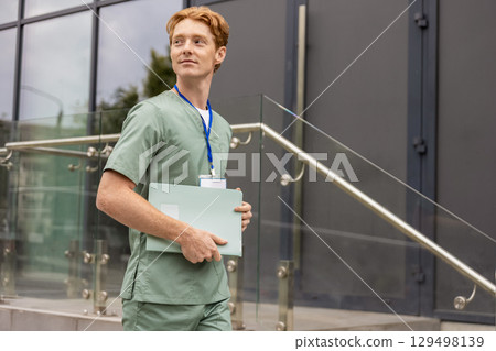 Young male doctor in medical scrubs walking hospital carrying clinical folder Young male doctor in medical scrubs walking hospital carrying clinical folder 129498139
