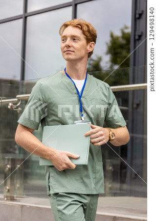 Red-haired healthcare worker in lab coat moving past glass building holding documents Red-haired healthcare worker in lab coat moving past glass building holding documents 129498140