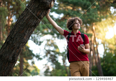 Curly-haired teen in red tshirt standing near the tree at the park 129498467