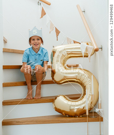 A cheerful five-year-old boy sits on the stairs with a big golden balloon, celebrating his birthday at home with festive decor 129499468