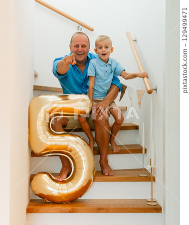 Candid birthday portrait of dad and son enjoying the moment on a staircase decorated with a festive balloon and garland Candid birthday portrait of dad and son enjoying the moment on a staircase decorated with a festive balloon and garland 129499471