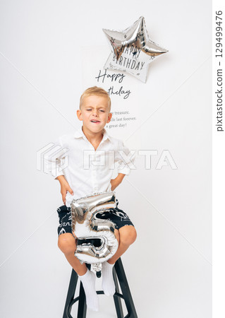Portrait of a young boy celebrating his 5th birthday, sitting on a stool with a silver number 5 balloon. Perfect for birthday themes, childhood, parenting, and celebration concepts. Portrait of a young boy celebrating his 5th birthday, sitting on a stool with a silver number 5 balloon. Perfect for birthday themes, childhood, parenting, and celebration concepts. 129499476