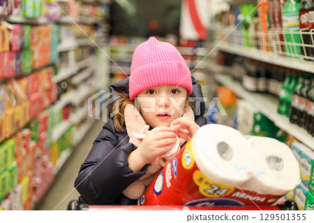 A young child wearing a pink hat is sitting in a shopping cart in a grocery store aisle, looking forward while holding something A young child wearing a pink hat is sitting in a shopping cart in a grocery store aisle, looking forward while holding something 129501355