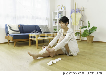A young woman sitting on the floor in the living room and putting on socks 129501786