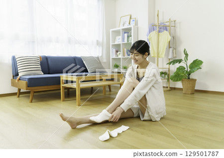 A young woman sitting on the floor in the living room and putting on socks 129501787