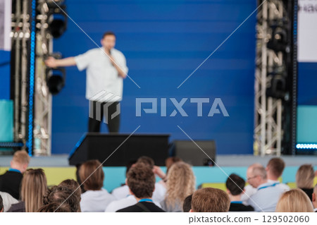 Speaker presenting to audience at professional event with blue stage backdrop 129502060