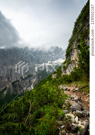 Mountain trail in the Dolomites (Dolomiti, Dolomiten), Italy, leading to Lago Sorapis. The path winds along rocky slopes with stunning views of valleys, forests, and peaks Mountain trail in the Dolomites (Dolomiti, Dolomiten), Italy, leading to Lago Sorapis. The path winds along rocky slopes with stunning views of valleys, forests, and peaks 129502187