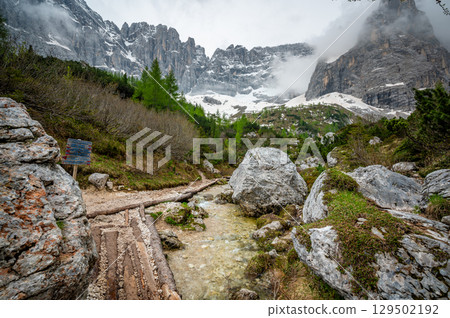 Rocky hiking trail near Lago Sorapis in the Dolomites (Dolomiti, Dolomiten), Italy. The path runs alongside a small mountain stream, with massive boulders and peaks in the background Rocky hiking trail near Lago Sorapis in the Dolomites (Dolomiti, Dolomiten), Italy. The path runs alongside a small mountain stream, with massive boulders and peaks in the background 129502192