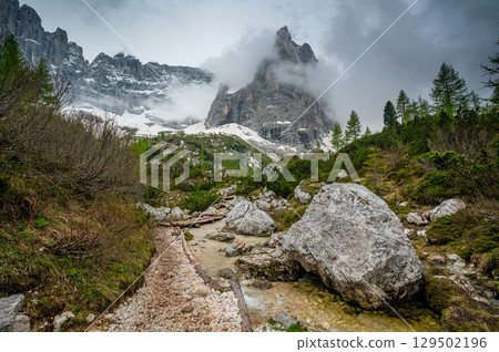 Rocky hiking trail near Lago Sorapis in the Dolomites (Dolomiti, Dolomiten), Italy. The path runs alongside a small mountain stream, with massive boulders and peaks in the background 129502196