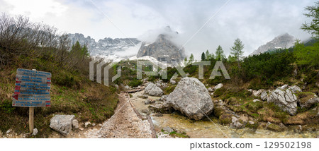 Rocky hiking trail near Lago Sorapis in the Dolomites (Dolomiti, Dolomiten), Italy. The path runs alongside a small mountain stream, with massive boulders and peaks in the background 129502198