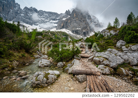 Rocky hiking trail near Lago Sorapis in the Dolomites (Dolomiti, Dolomiten), Italy. The path runs alongside a small mountain stream, with massive boulders and peaks in the background Rocky hiking trail near Lago Sorapis in the Dolomites (Dolomiti, Dolomiten), Italy. The path runs alongside a small mountain stream, with massive boulders and peaks in the background 129502202