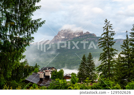Scenic mountain village in the Dolomites (Dolomiti, Dolomiten), Italy. Cozy alpine houses on green slopes, towering peaks above, and clouds rolling through the valley below 129502626