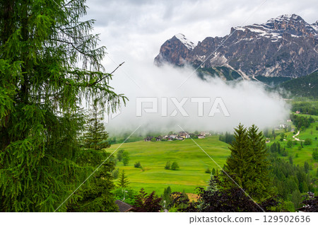 Scenic mountain village in the Dolomites (Dolomiti, Dolomiten), Italy. Cozy alpine houses on green slopes, towering peaks above, and clouds rolling through the valley below Scenic mountain village in the Dolomites (Dolomiti, Dolomiten), Italy. Cozy alpine houses on green slopes, towering peaks above, and clouds rolling through the valley below 129502636