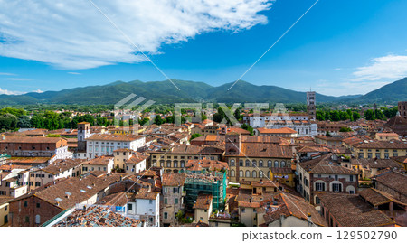 Panoramic view of Lucca, Italy, with red brick rooftops and historic churches seen from above. Tuscan hills and mountains form a stunning summer backdrop under a clear blue sky. 129502790