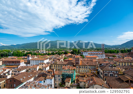 Panoramic view of Lucca, Italy, with red brick rooftops and historic churches seen from above. Tuscan hills and mountains form a stunning summer backdrop under a clear blue sky. 129502791