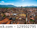 Panoramic view of Lucca, Italy, with red brick rooftops and historic churches seen from above. Tuscan hills and mountains form a stunning summer backdrop under a clear blue sky. 129502795