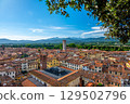 Panoramic view of Lucca, Italy, with red brick rooftops and historic churches seen from above. Tuscan hills and mountains form a stunning summer backdrop under a clear blue sky. 129502796