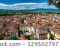 Panoramic view of Lucca, Italy, with red brick rooftops and historic churches seen from above. Tuscan hills and mountains form a stunning summer backdrop under a clear blue sky. 129502797