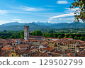 Panoramic view of Lucca, Italy, with red brick rooftops and historic churches seen from above. Tuscan hills and mountains form a stunning summer backdrop under a clear blue sky. 129502799