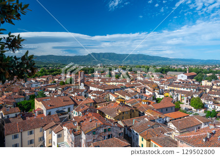 Panoramic view of Lucca, Italy, with red brick rooftops and historic churches seen from above. Tuscan hills and mountains form a stunning summer backdrop under a clear blue sky. 129502800