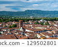 Panoramic view of Lucca, Italy, with red brick rooftops and historic churches seen from above. Tuscan hills and mountains form a stunning summer backdrop under a clear blue sky. 129502801