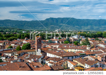 Panoramic view of Lucca, Italy, with red brick rooftops and historic churches seen from above. Tuscan hills and mountains form a stunning summer backdrop under a clear blue sky. Panoramic view of Lucca, Italy, with red brick rooftops and historic churches seen from above. Tuscan hills and mountains form a stunning summer backdrop under a clear blue sky. 129502801