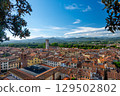 Panoramic view of Lucca, Italy, with red brick rooftops and historic churches seen from above. Tuscan hills and mountains form a stunning summer backdrop under a clear blue sky. 129502802